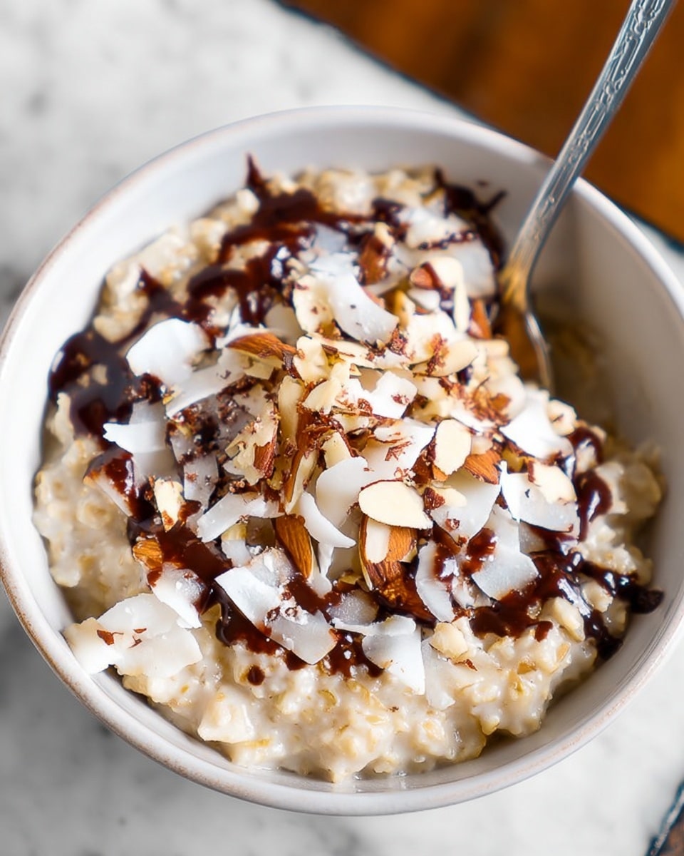 A close-up view of a creamy oatmeal bowl filled to the top with a thick, textured layer of warm, pale beige oats mixed with milk. Mixed within are thin, large white coconut flakes that add a delicate crunch, scattered alongside almond slices that show a light brown, toasted color. A generous drizzle of dark, glossy chocolate sauce is spread unevenly over the oats, adding rich contrast. The oatmeal bowl is smooth and white, set against a white marbled surface, with a shiny silver spoon placed inside on the right side, partially submerged in the oats. photo taken with an iphone --ar 4:5 --v 7