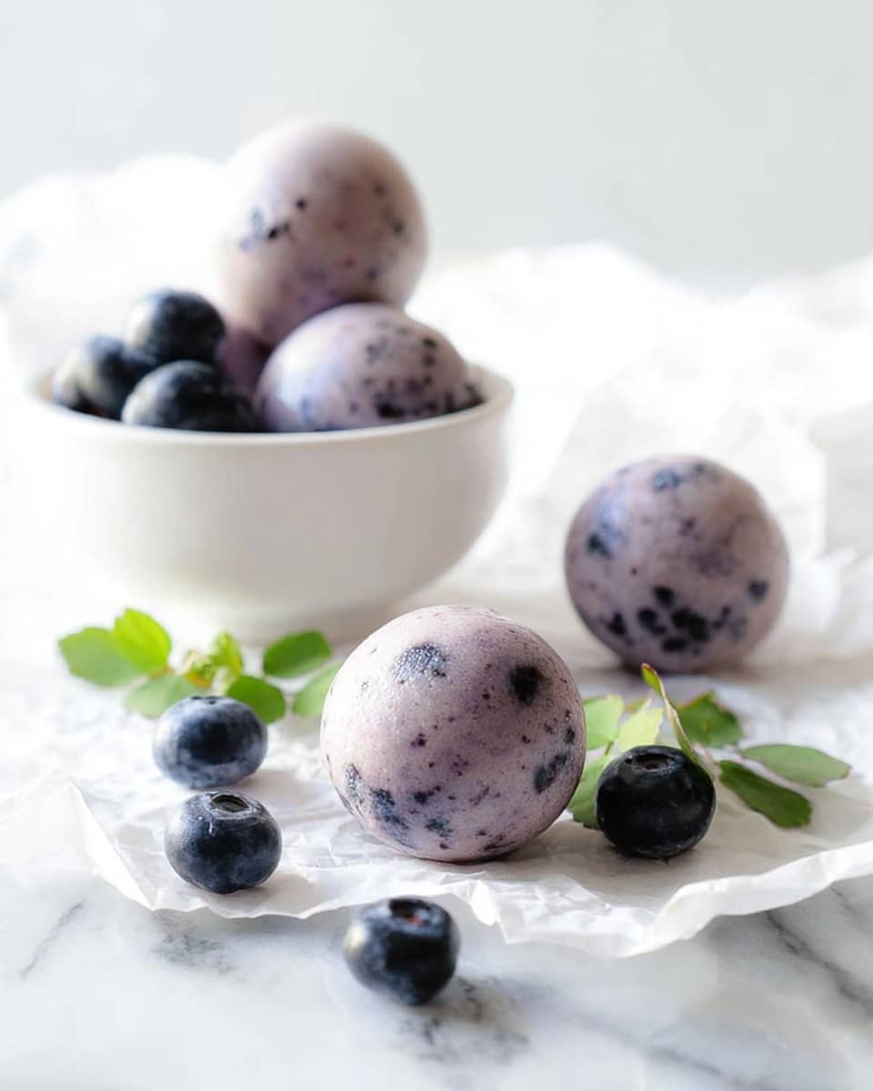 The image shows round, smooth balls that are light purple with small dark purple specks inside, arranged on a white sheet of crinkled paper on a white marbled surface. Around the balls are several fresh dark blue blueberries and two small green leaves for decoration. In the background, there is a white bowl holding two more of the same purple balls and several blueberries. The scene is bright and softly lit, making the colors look fresh and natural. photo taken with an iphone --ar 4:5 --v 7