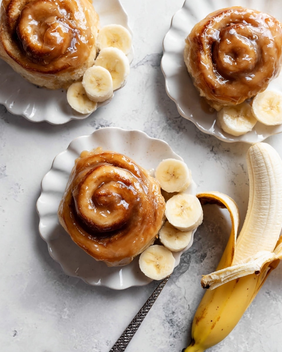 The image shows three sticky cinnamon rolls placed on white plates with a wavy edge, all set on a white marbled texture. Each cinnamon roll is a soft light brown swirl with thick, shiny caramel glaze covering the surface, adding a glossy texture. Around the cinnamon rolls on the plates are slices of ripe, pale yellow banana with soft, creamy centers. At the right side of the front plate, there is a partially peeled banana with its skin curling down and one round banana slice tucked inside, showing the fruit’s smooth texture. A silver spoon with a patterned handle rests next to the front cinnamon roll. Photo taken with an iphone --ar 4:5 --v 7