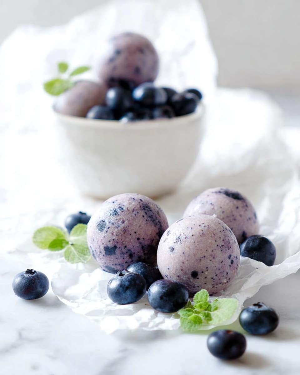Four light purple balls with dark specks, looking smooth and round, are placed on crumpled white paper in front of a white bowl. The bowl contains more of the same balls along with several fresh dark blue blueberries. Additional blueberries are scattered around the balls on the paper, accompanied by a few small green leaves. The whole scene rests on a white marbled surface, with soft natural light brightening the image. photo taken with an iphone --ar 4:5 --v 7