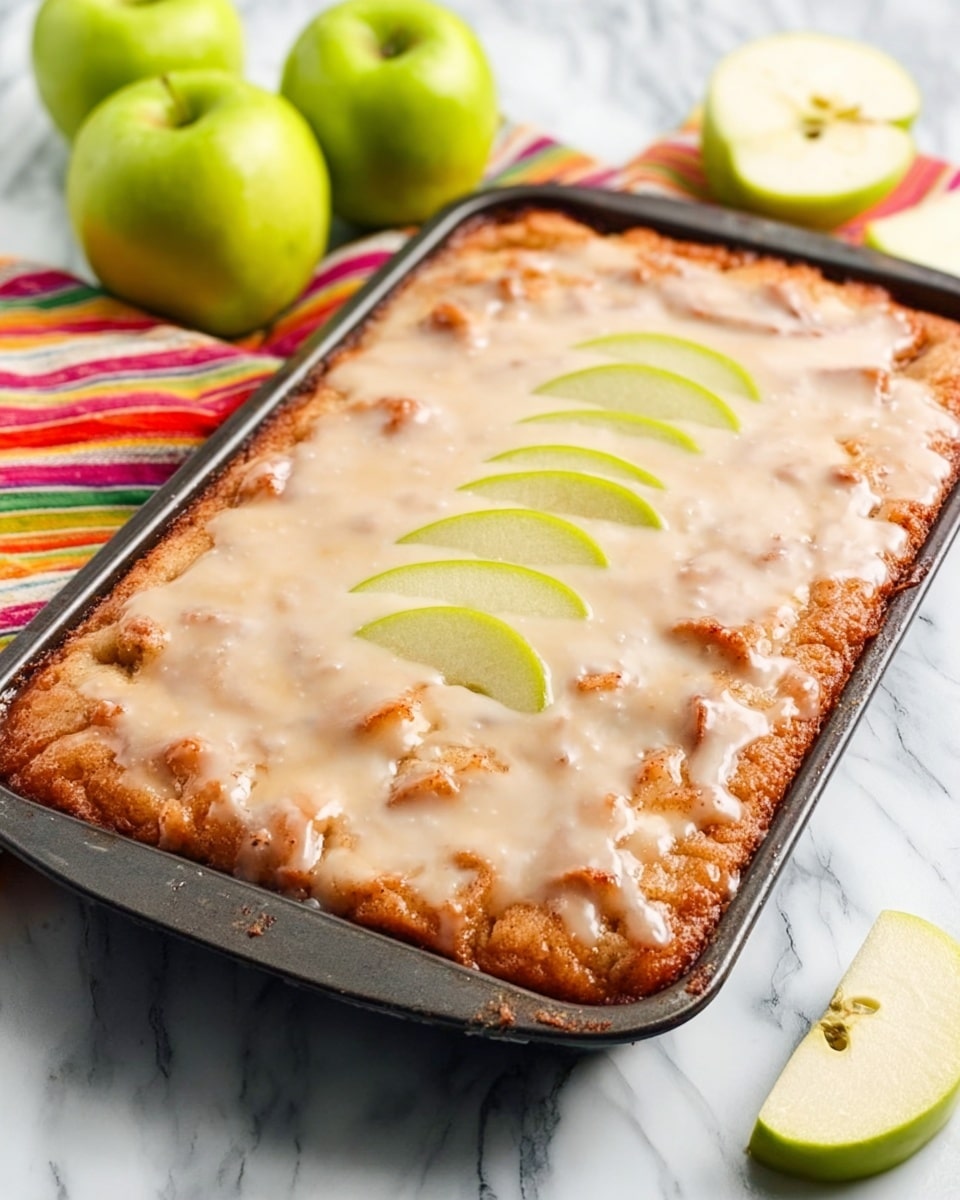 A rectangular baked apple dessert in a black metal pan with a light brown crust on the bottom and a thick, soft apple filling visible under a shiny, smooth white glaze covering the top. On the center of the dessert, there are five thin slices of green apple fanned out neatly. The pan is placed on a white marbled surface with three whole green apples, one cut in half showing the inside, and a folded colorful striped cloth in the background. photo taken with an iphone --ar 4:5 --v 7