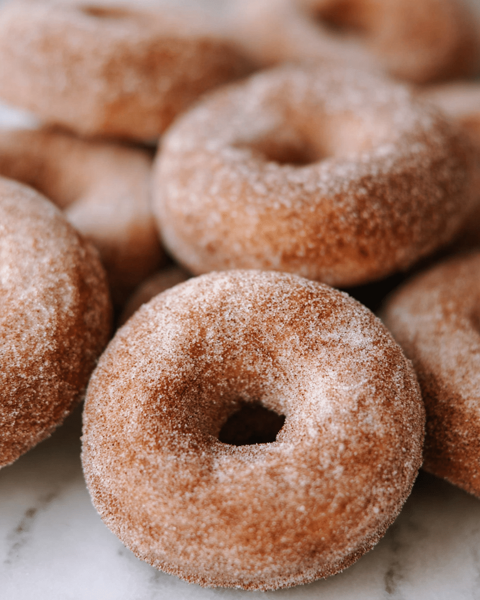 The image shows a close-up of many round cinnamon sugar donuts placed closely together on a white marbled surface. Each donut has a light brown color with a slightly rough texture from the sugar coating, and a small hole in the center. The donuts look soft and have an even dusting of sugar and cinnamon, giving them a grainy and matte finish. The focus is on the front donut, with the others blurring softly into the background. Photo taken with an iphone --ar 4:5 --v 7