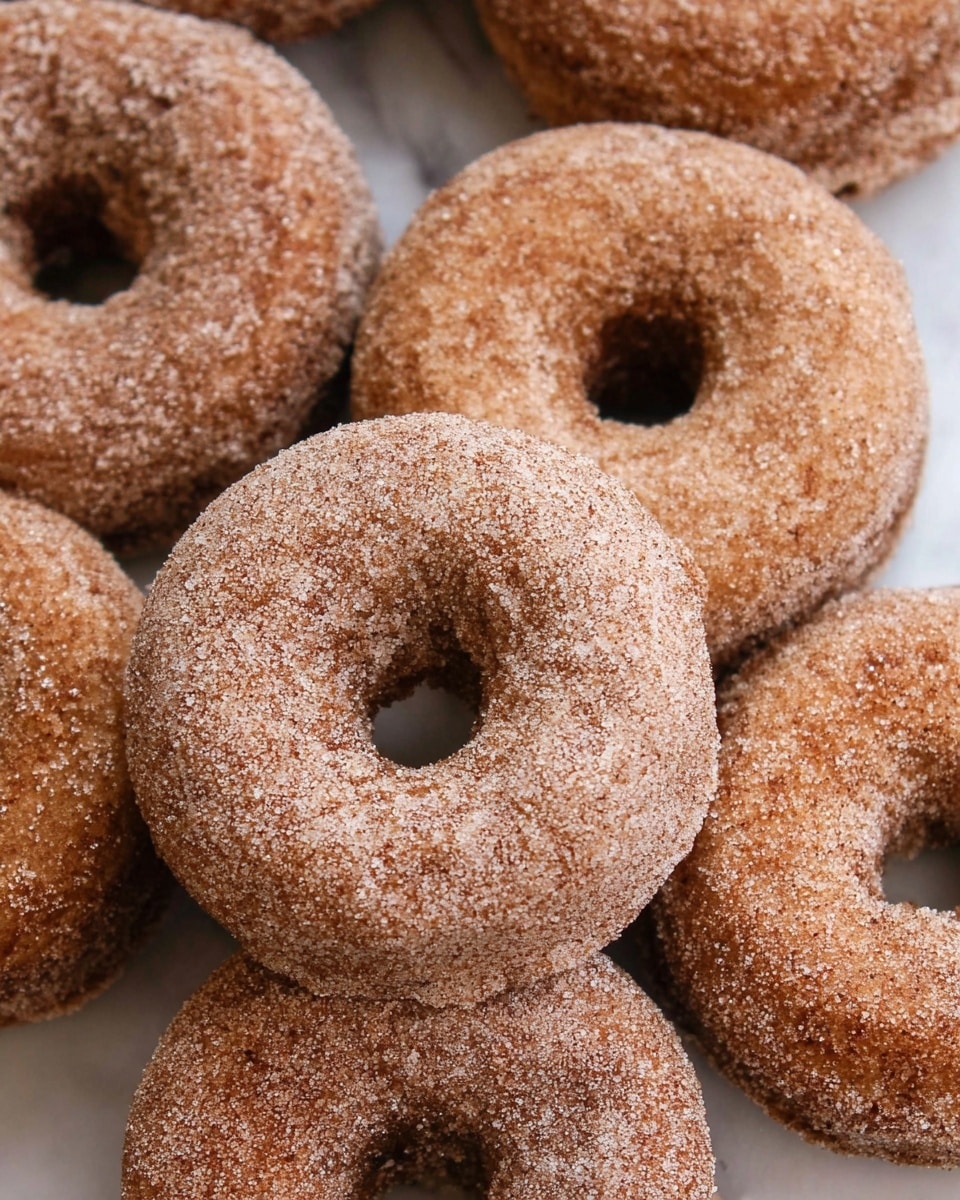The image shows a close-up of several cinnamon sugar-coated donuts arranged closely together. Each donut is round with a hole in the middle and has a rough texture from the sugar and cinnamon coating, giving them a light brown color with specks of white sugar crystals all over. The donuts appear to be soft, puffy, and evenly covered, placed on a white marbled surface. photo taken with an iphone --ar 4:5 --v 7