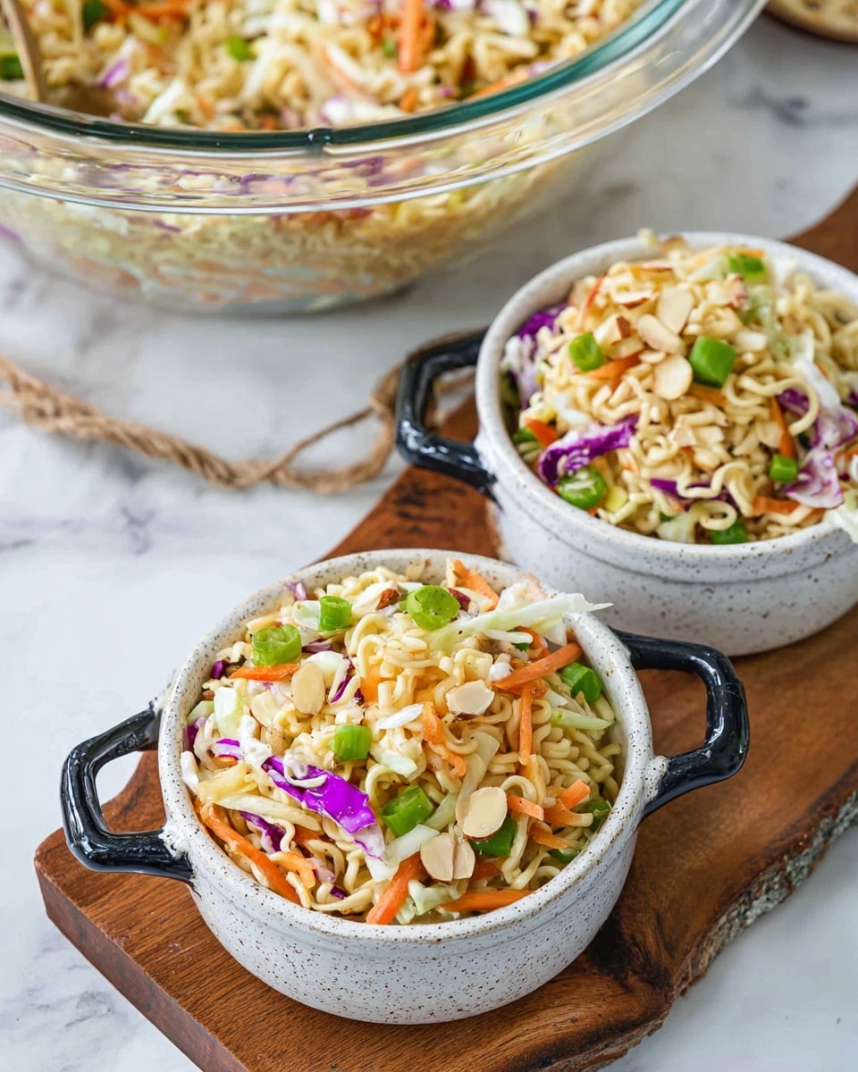 The image shows two small white speckled bowls with black handles, each filled with a colorful noodle salad. The salad has three main layers: a base of curly, light beige ramen noodles, topped with shredded cabbage in white and purple, thin orange carrot strips, chopped green onions, and sliced almonds scattered throughout. The bowls are placed on a wooden board with a white marbled surface underneath. In the background, there is a large clear glass bowl filled with the same salad. The textures range from soft noodles to crunchy nuts and fresh vegetables. Photo taken with an iphone --ar 4:5 --v 7