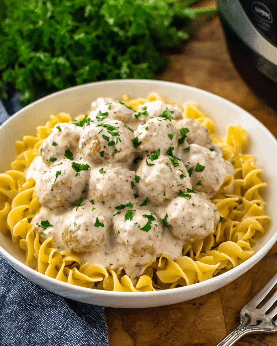 A white bowl filled with three layers: the bottom layer is yellow curly egg noodles covering the full bowl; on top of the noodles, there is a thick layer of round meatballs coated in a creamy white sauce with pepper specks; the top layer is garnished with small green parsley pieces scattered across the sauce and meatballs. The bowl is set on a wooden surface with a silver fork beside it, and a bunch of green parsley is blurred in the background. Photo taken with an iphone --ar 4:5 --v 7