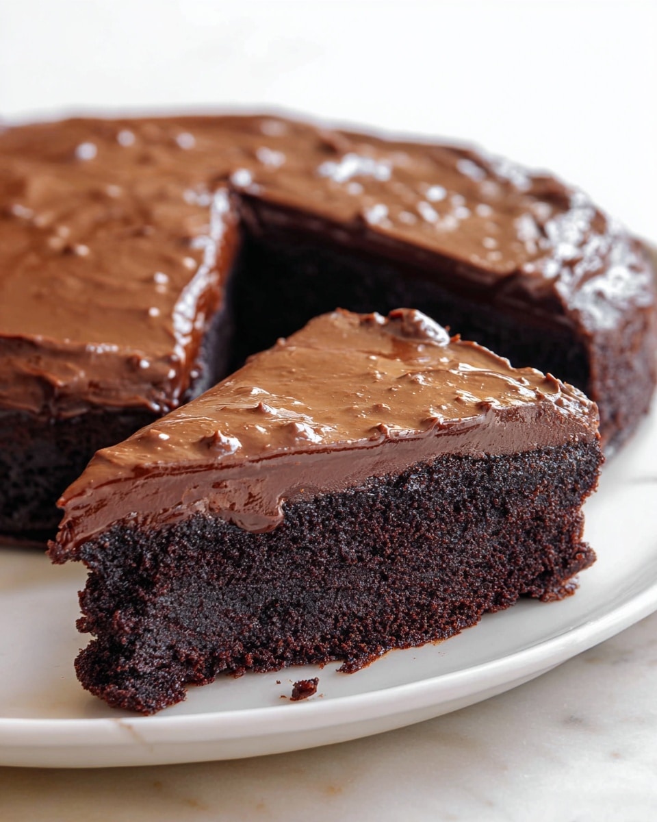 A rich, dense chocolate cake slice with one thick layer that is dark brown and moist, topped with a thick, glossy, slightly textured milk chocolate frosting covering the top fully with small uneven bumps. The slice is lifted slightly above a whole round cake of the same type, set on a white plate on a white marbled surface. The edges of the frosting drip slightly over the sides of the cake, showing the dense texture below. photo taken with an iphone --ar 4:5 --v 7