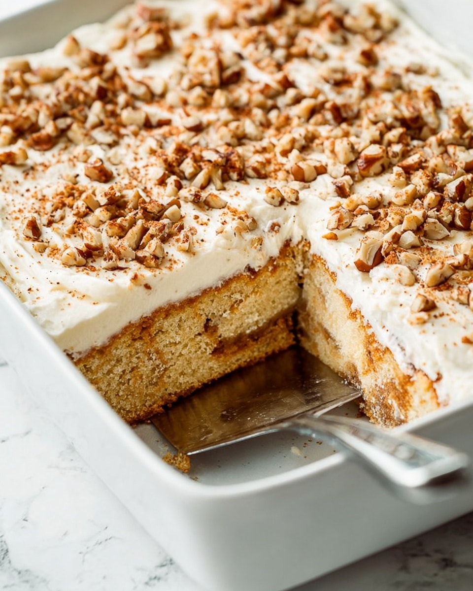 A close-up of a square white baking dish filled with a layered cake that has a light golden spongy base with a visible cinnamon swirl in the middle, topped with a thick layer of creamy white frosting, generously sprinkled with chopped nuts that have a light brown and tan color. One large square piece is cut out, showing the soft texture of the cake inside and a metal spatula underneath the cut piece. The background is a white marbled surface. photo taken with an iphone --ar 4:5 --v 7