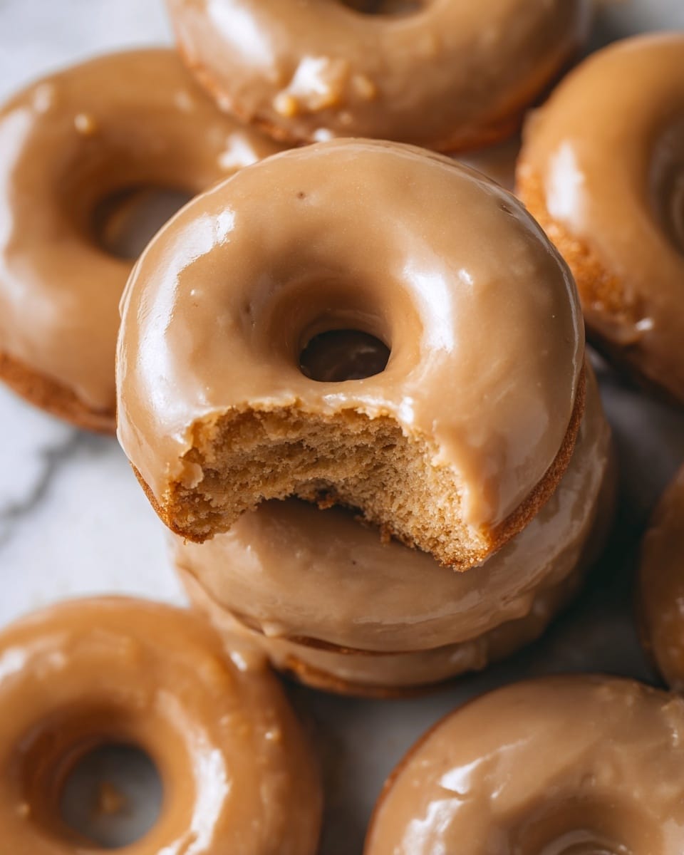 The image shows a stack of three donuts with a light brown glaze on top, placed on white parchment paper resting on a wooden board. The top donut is broken into pieces, revealing a soft, porous, light brown inside texture. The glaze on the donuts is smooth and slightly cracked, dripping a bit on the edges but covering the top fully. The white marbled texture below adds clean contrast to the warm colors of the donuts and board. photo taken with an iphone --ar 4:5 --v 7