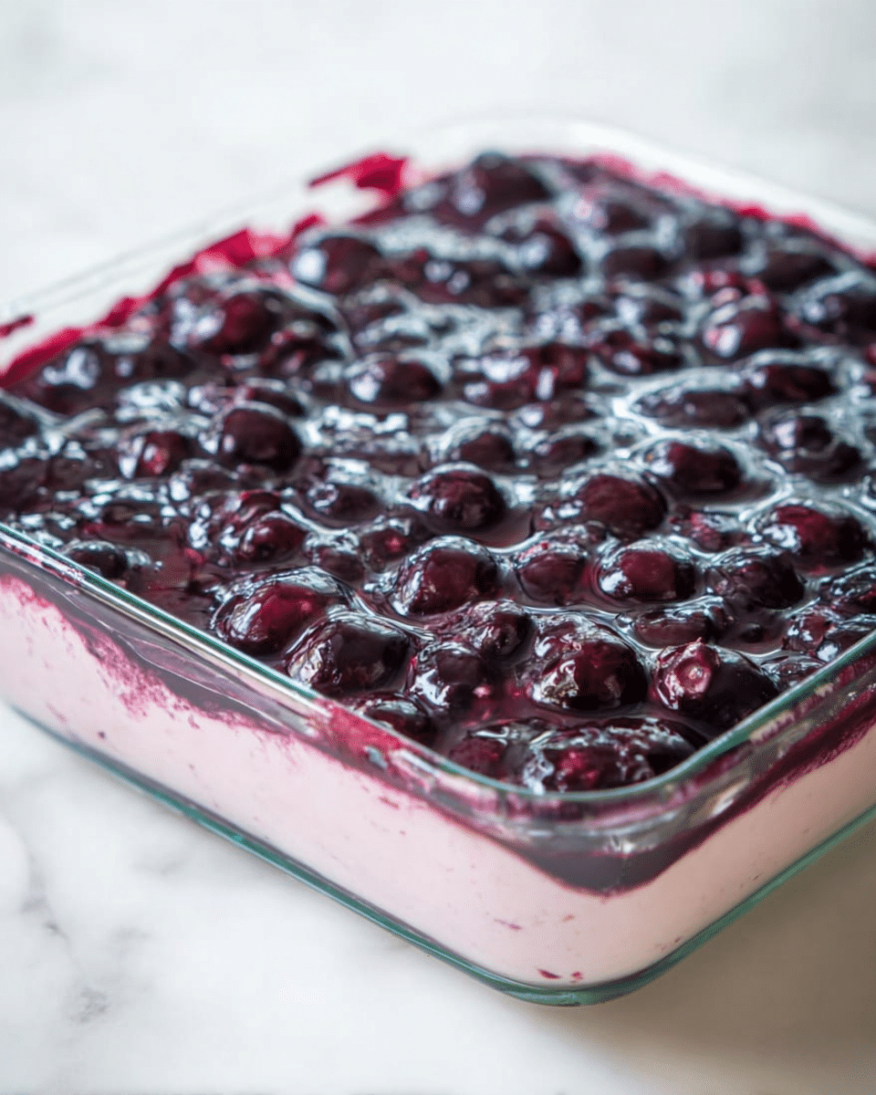 The image shows a close-up of a square glass dish filled with a dessert that has two visible layers; the bottom layer is a smooth, pale pink cream, and the top layer is a thick, glossy dark purple-blue sauce with whole blueberries embedded throughout. The fruit layer looks juicy and shiny, contrasting with the creamy layer underneath. The dish is placed on a white marbled surface. photo taken with an iphone --ar 4:5 --v 7
