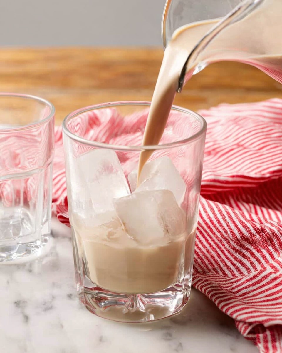A clear glass pitcher filled almost to the top with a smooth, creamy beige liquid. The pitcher has a curved handle on the right side and a very small pointed spout on the left. It is placed on a white marbled surface with a red and white striped cloth casually draped behind it. The wood grain floor adds a warm background. The texture of the liquid looks thick and rich, filling the pitcher evenly without bubbles. photo taken with an iphone --ar 4:5 --v 7