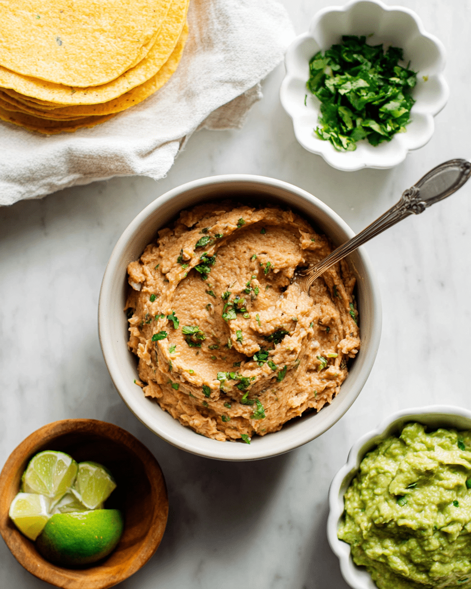 The image shows a white bowl filled with light brown refried beans, mixed smoothly with some green herbs sprinkled on top, giving a soft textured look. A silver spoon rests inside the bowl, partially covered by the beans. To the upper right, there is a white bowl containing bright green guacamole with a creamy texture. Above the main bowl is a small white scalloped dish holding fresh chopped cilantro. In the upper left corner, there are some yellow corn tortillas stacked on a white cloth. To the left of the bowl with beans, there is a small wooden bowl holding lime wedges. All these items sit on a white marbled surface. photo taken with an iphone --ar 4:5 --v 7