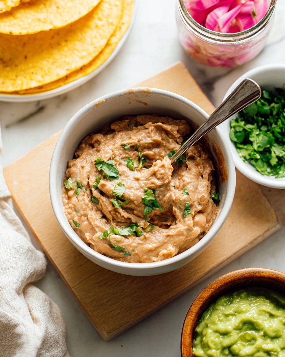 A close-up of a white bowl filled with creamy, light brown refried beans topped with pieces of green cilantro; the beans have a slightly chunky texture with a spoon resting inside the bowl. Around the bowl, there is a white small dish with chopped cilantro, a wooden bowl with a green sauce, a glass jar of pink pickled onions, and a white bowl holding a smooth, bright green guacamole or avocado dip. To the left, part of a white cloth and yellow corn tortillas appear on a white marbled surface, all set on a light wooden board. Photo taken with an iphone --ar 4:5 --v 7