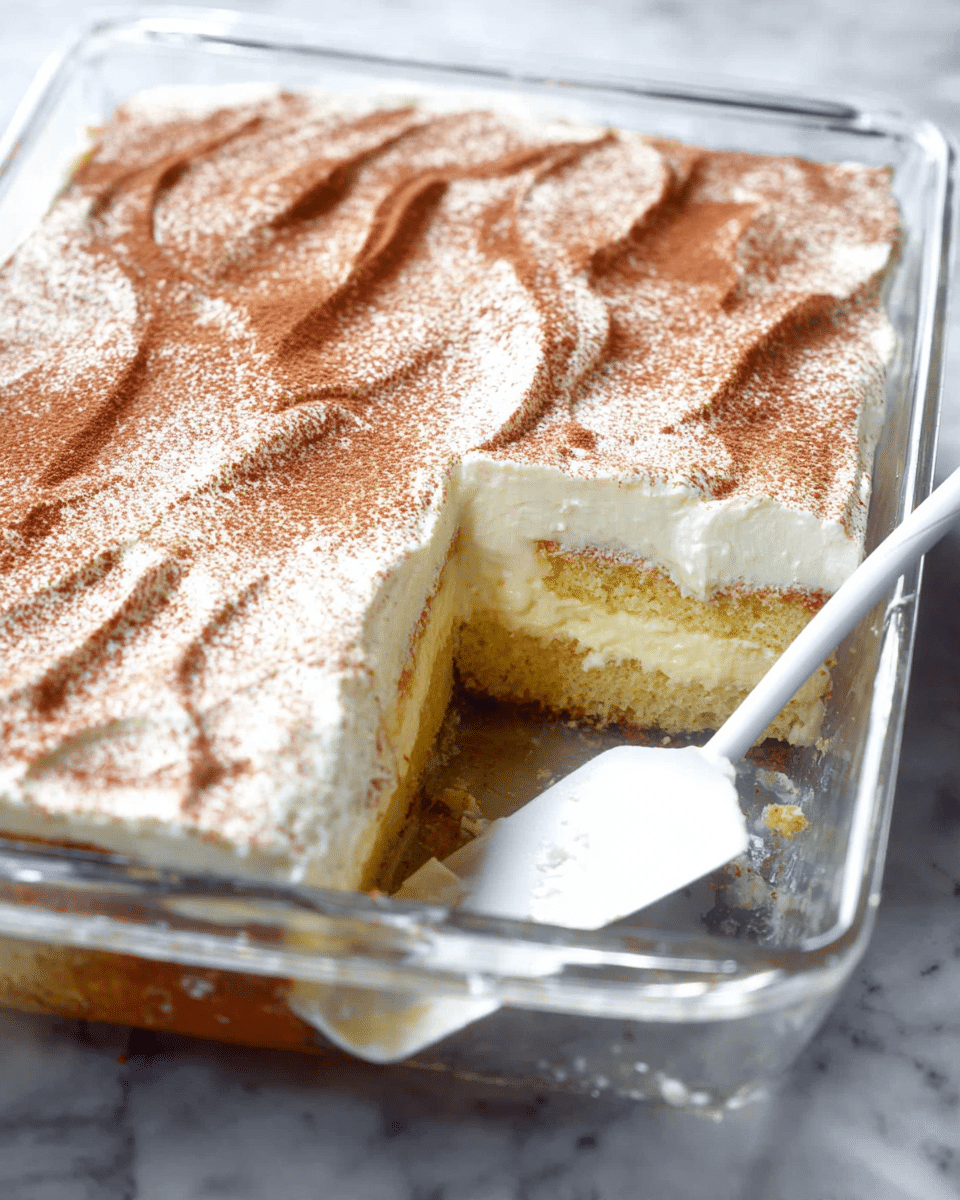 A square piece of cake with two main layers sits on a white plate on a white marbled texture. The bottom layer is a moist, light yellow sponge cake with small air holes visible, while the top layer is a thick, white creamy frosting dusted with a light sprinkle of brown cinnamon powder. On top, a red strawberry, sliced thinly and fanned out, adds a bright splash of color. To the side, a silver fork rests on the plate, and in the background, a gray cloth and another strawberry are slightly blurred. Photo taken with an iphone --ar 4:5 --v 7