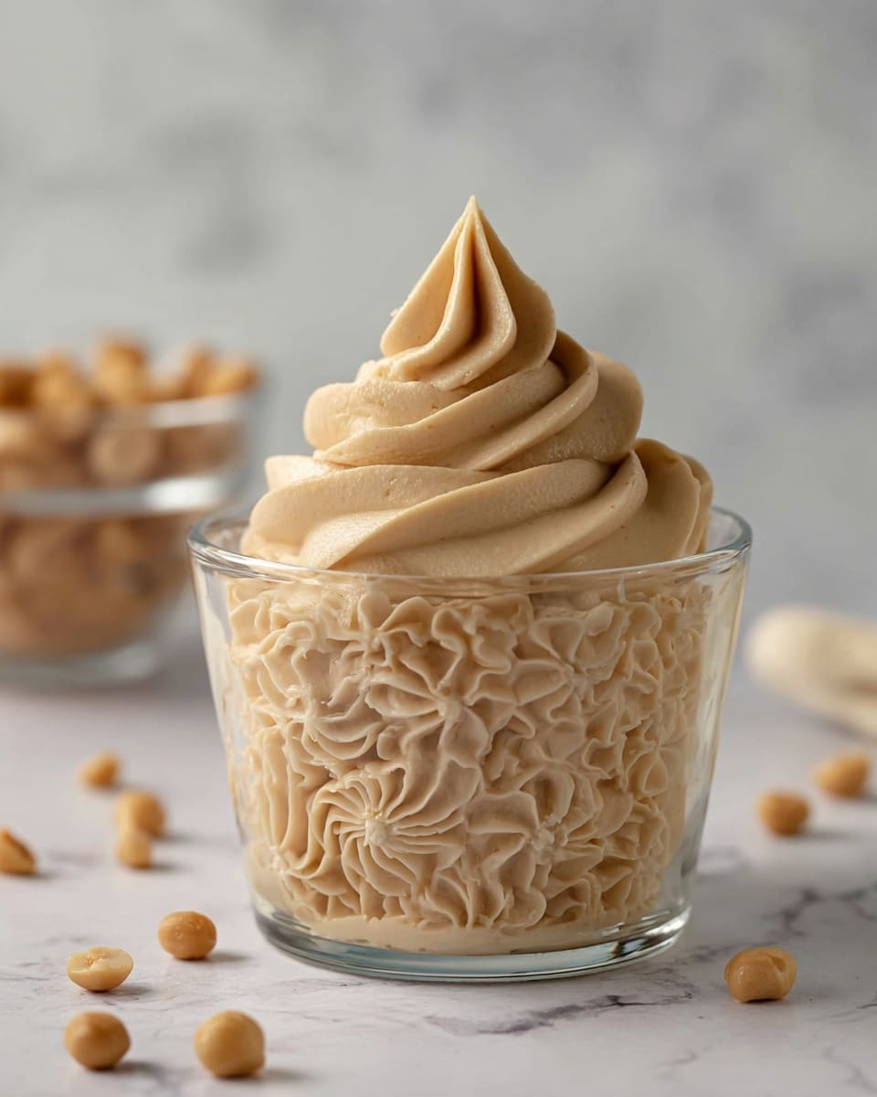 A clear glass cup is filled with creamy, light brown frosting in two layers: the bottom layer has small, intricate rosettes and swirls covering the entire inside of the cup, and the top layer is a smooth, tall swirl with a pointed tip rising above the rim. Scattered around the cup are small round light brown nuts, and the background shows a blurred glass bowl on a white marbled texture. photo taken with an iphone --ar 4:5 --v 7