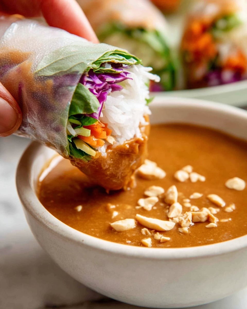 A close-up of a fresh spring roll being held by a woman's hand, dipped halfway into a bowl of thick, light brown peanut sauce with small chopped peanuts on the surface. The spring roll has a translucent white rice paper wrap showing the layers inside: white rice noodles at the top, purple shredded cabbage, green fresh herbs, and orange shredded carrots, all tightly packed. The bowl is white and sits on a white marbled surface that is out of focus in the background. Photo taken with an iphone --ar 4:5 --v 7