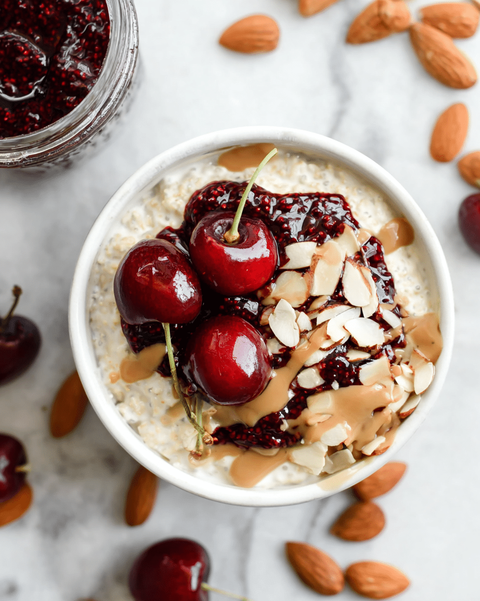 A white bowl filled with creamy oatmeal forms the base layer, topped with a dark red cherry chia jam spread unevenly over one side. Two shiny, fresh cherries with stems rest on top of the jam, adding a bright red pop of color. Light brown almond butter is drizzled generously over the oatmeal and jam, with thinly sliced almonds scattered around, adding texture and contrast. The bowl is on a white marbled surface, and nearby whole and sliced almonds are visible, along with a small jar containing similar oats and toppings. photo taken with an iphone --ar 4:5 --v 7