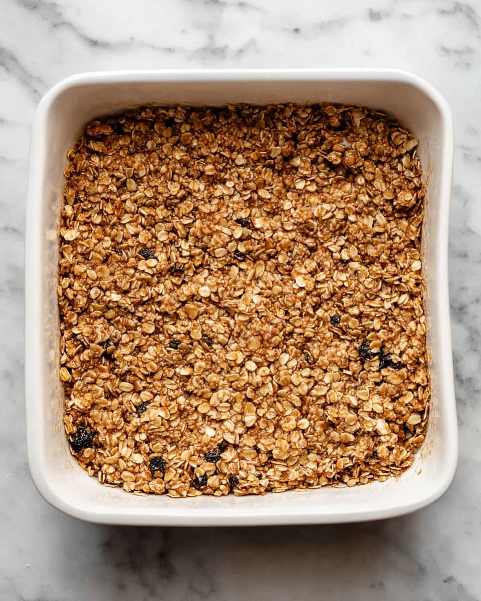 A square white casserole dish filled with one thick layer of golden-brown baked oat mixture that looks crunchy and textured with visible rolled oats and small dark bits, placed on a white marbled surface, photo taken with an iphone --ar 4:5 --v 7