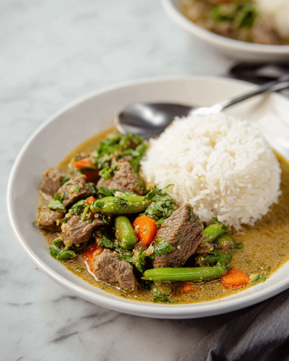 The image shows a white plate with two main layers: on the right, a dome-shaped mound of white steamed rice with a fluffy texture, and on the left, a thick curry filled with tender brown beef slices, bright green pea pods, orange carrot pieces, and fresh green herbs scattered throughout. The curry has a light brown sauce with a slightly oily surface, pooling around the rice and beef. A silver spoon rests behind the rice inside the plate. The dish sits on a white marbled surface with soft natural lighting. Photo taken with an iphone --ar 4:5 --v 7