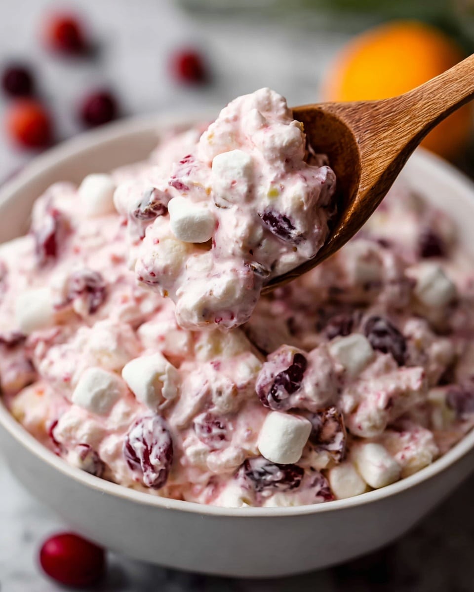A white bowl filled with a creamy pink mixture that has many small white marshmallows and tiny red fruit pieces spread evenly throughout. The texture looks thick and slightly chunky with small bits of darker red and orange fruits mixed in. Around the bowl, there are a few whole cranberries, mini marshmallows, and bright orange slices placed on a white marbled surface. A wooden spoon with a smooth dark brown handle is positioned near the bowl. Photo taken with an iphone --ar 4:5 --v 7