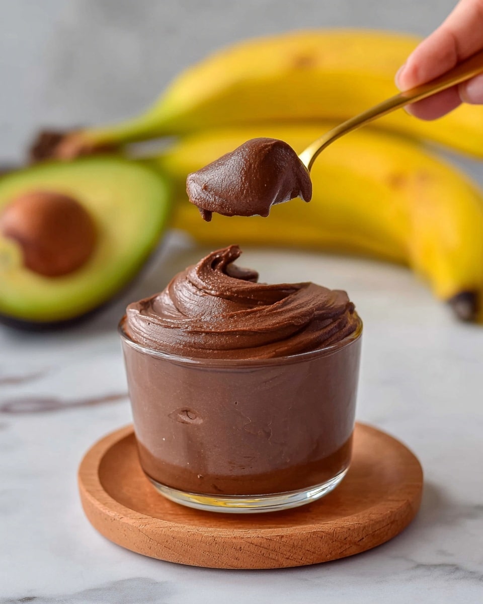 A small transparent glass cup filled with smooth, dark brown chocolate mousse, slightly overflowing at the top, resting on a round wooden coaster placed on a white marbled surface; a woman's hand holds a small golden spoon lifting a large, fluffy dollop of the mousse above the cup; bright yellow bananas and a halved avocado with its large brown seed sit blurred in the background. photo taken with an iphone --ar 4:5 --v 7