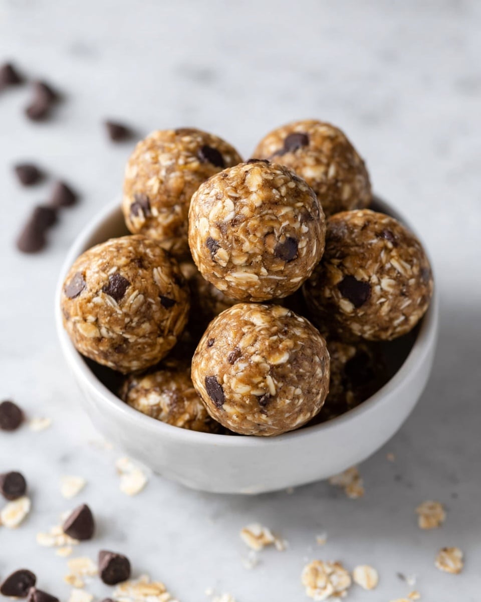 A white bowl filled with eight round energy balls made of oats and chocolate chips. The balls have a rough texture from the oats and small, dark chocolate chips scattered throughout. Some extra chocolate chips are scattered around the bowl on a white marbled surface. The image focuses closely on the bowl and the energy balls, showing the detailed texture and colors of the ingredients, with warm natural light highlighting the oat flakes and chocolate pieces. photo taken with an iphone --ar 4:5 --v 7