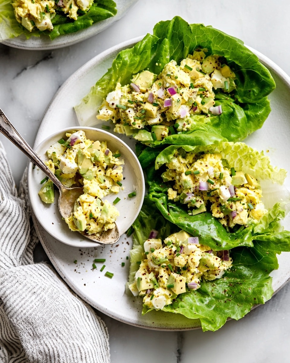 A white plate sits on a white marbled surface, holding three lettuce wraps with bright green, ruffled lettuce leaves as the base layer. Each leaf is filled with a chunky mix of pale yellow and white chopped boiled eggs, light green chunks of avocado, small pieces of red onion, and sprinkled with finely chopped chives and black pepper, creating a textured and colorful look. A small white bowl filled with the same egg and avocado mixture sits on the plate with a spoon inside it. A white and gray striped cloth is partially visible in the bottom left corner. Photo taken with an iphone --ar 4:5 --v 7