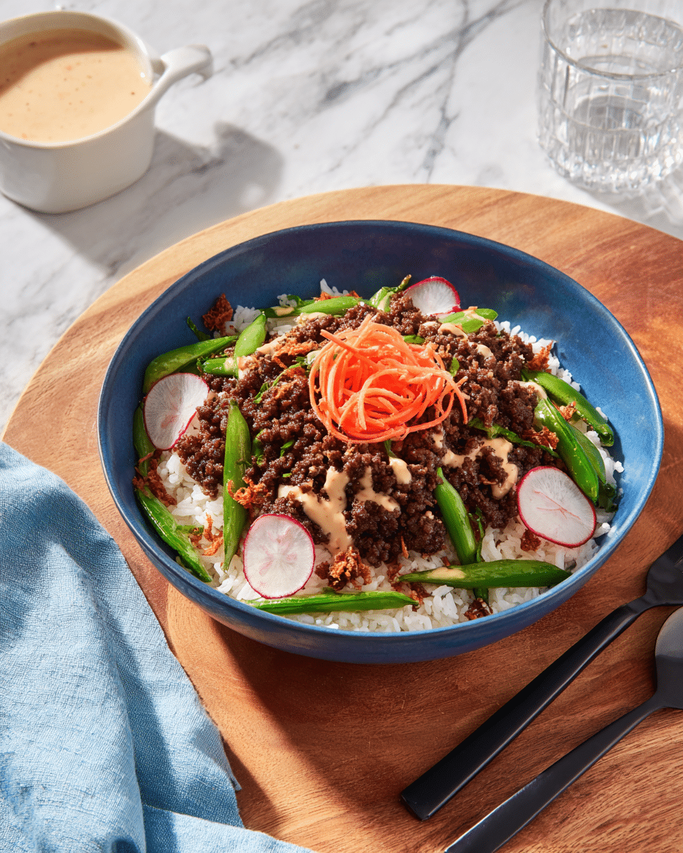 A blue bowl filled with a base layer of white fluffy rice, topped with a layer of cooked ground beef that is dark brown in color and scattered evenly over the rice. On top of the beef are fresh green snap peas, thinly sliced round radish pieces with white centers and pink edges, and a small mound of thinly sliced orange carrot strips in the center. Light beige sauce is drizzled over the entire dish, and small crispy brown bits are sprinkled across the top. The bowl sits on a wooden board with a pale blue cloth napkin to the left, a black fork to the right, a small white bowl with creamy beige sauce above, and a glass to the upper right, all placed on a white marbled texture surface. Photo taken with an iphone --ar 4:5 --v 7