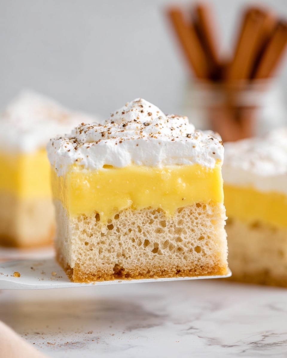 The image shows a close-up of a three-layer square piece of cake held by a white spatula, set against a white marbled surface. The bottom layer is a light brown sponge cake with a soft, porous texture and small visible holes. Above that is a thick, smooth, bright yellow custard layer with a creamy look. The top layer is a fluffy white whipped cream layer, evenly spread and slightly textured with small brown specks sprinkled on top. In the background, blurred, are more similar cake squares and cinnamon sticks in a glass jar. photo taken with an iphone --ar 4:5 --v 7