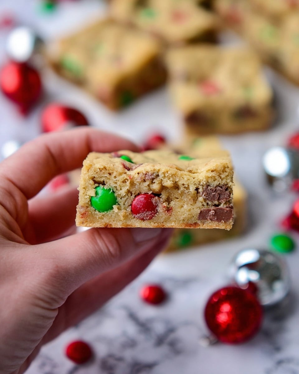 A close-up view of a square blondie bar held between a woman's thumb and forefinger, showing one layer with golden brown crumbly texture mixed with colorful candy-coated chocolate pieces in red and green. In the blurred background, more blondie squares lie flat on a white marbled surface, decorated with scattered red and green candies and shiny silver Christmas ornaments hinting a festive theme. The image has soft lighting that highlights the texture and colors, focusing sharply on the blondie piece in the foreground. Photo taken with an iphone --ar 4:5 --v 7
