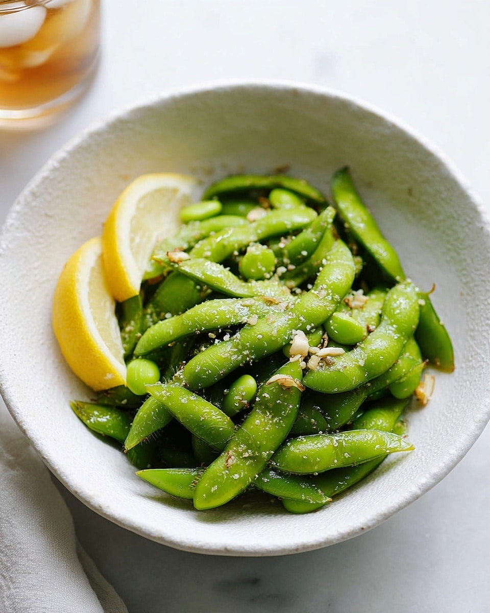 A white textured bowl filled with bright green edamame pods sprinkled with coarse salt and small garlic pieces, showing a slightly shiny and fresh texture. Two lemon wedges with a pale yellow color rest on the left side of the bowl, adding a fresh contrast. The bowl sits on a white marbled surface with soft natural light highlighting the vivid colors and textures inside. Part of a glass with iced drink is visible in the top left corner. photo taken with an iphone --ar 4:5 --v 7