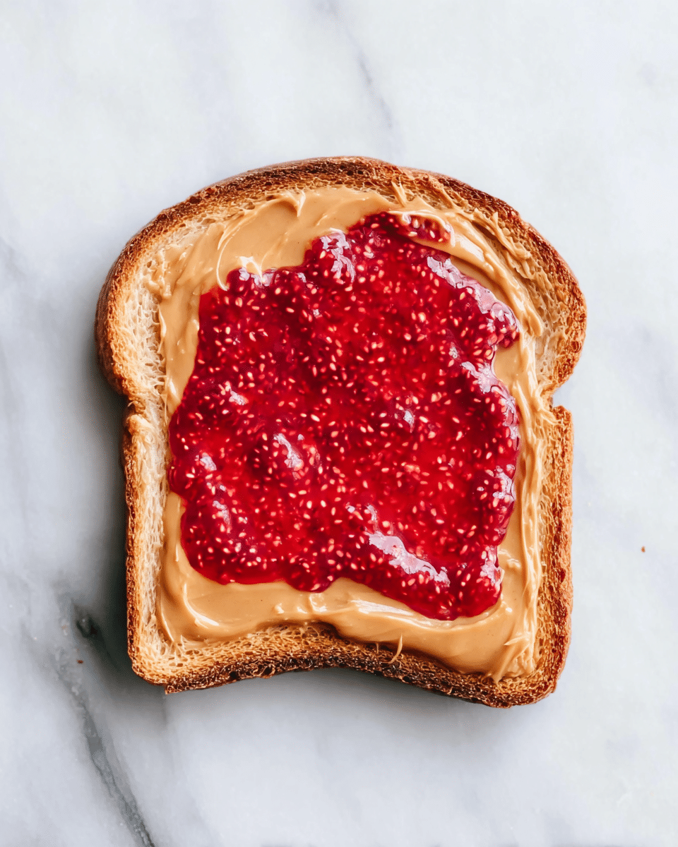 A single slice of toasted bread lies flat on a white marbled surface. The toast is golden-brown with slightly darker edges showing a crunchy texture. On top, there are two distinct layers: the bottom layer is a smooth, creamy light brown spread of peanut butter, evenly covering most of the slice but leaving a thin border of bread exposed. The top layer is a thick, glossy bright red raspberry jam with visible seeds, spread roughly in the center but not covering the entire peanut butter layer. The contrast between the creamy peanut butter and the chunky, textured raspberry jam is clear. Photo taken with an iphone --ar 4:5 --v 7