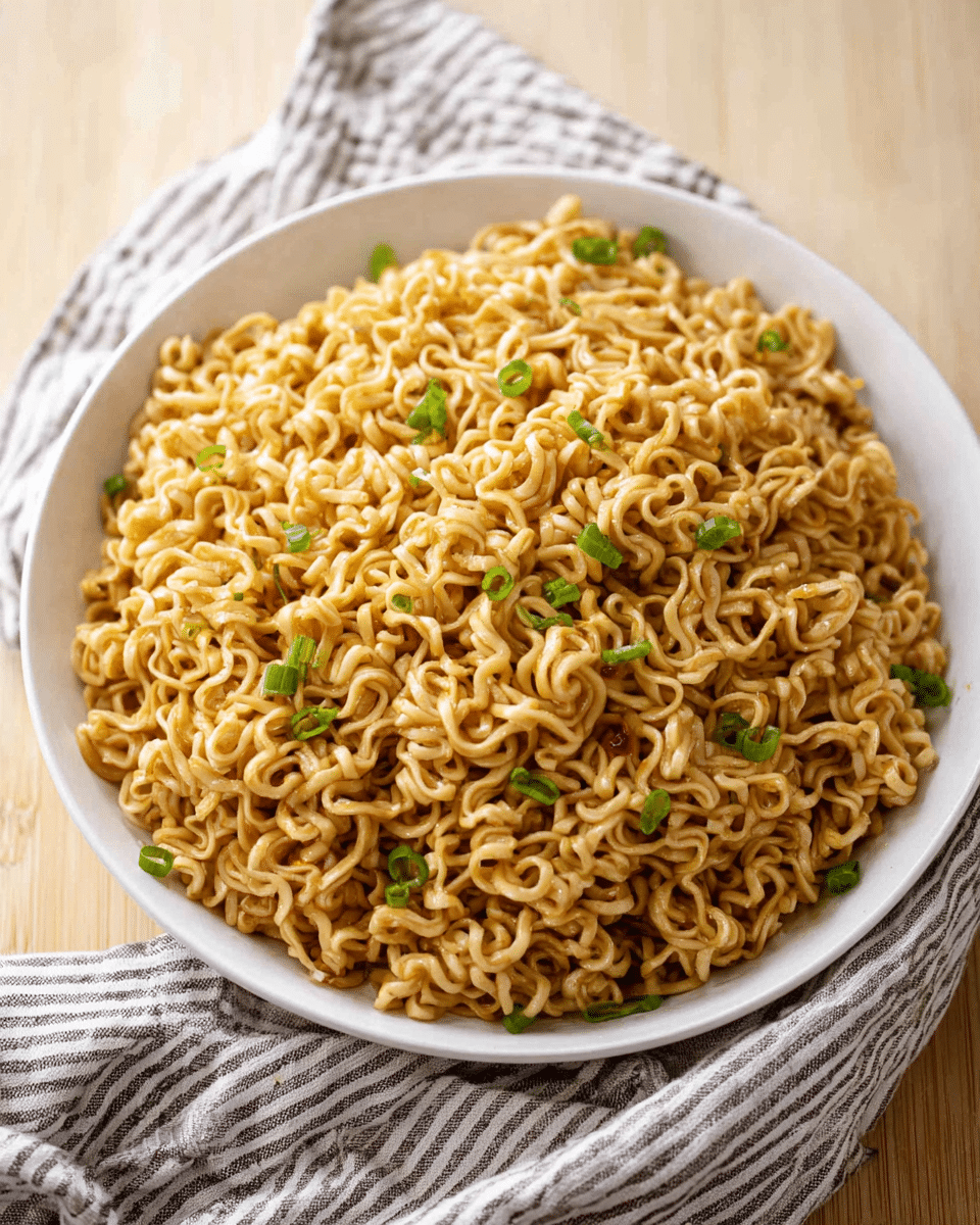 A white bowl filled with a large serving of cooked instant noodles, the noodles have a light brown color and a slightly glossy, oily texture. The noodles are piled high with some loose strands falling naturally over the edges. Small pieces of bright green chopped scallions are scattered evenly on top, adding a fresh contrast. The bowl is placed on a light wooden surface with a striped cloth in the background. photo taken with an iphone --ar 4:5 --v 7