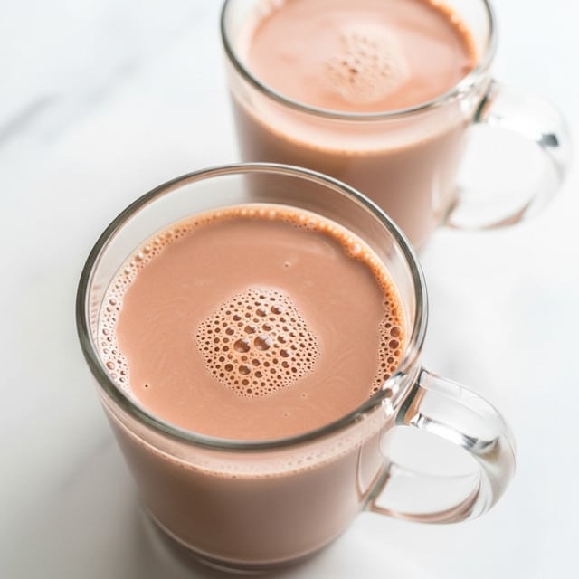 A clear glass mug filled with rich, creamy hot chocolate with a smooth light to medium brown color and small bubbles on the surface, showing a velvety texture. Another mug filled with the same hot chocolate is slightly blurred in the background to the upper right, both placed on a white marbled surface. The glass mugs have thick handles and the hot chocolate fills almost to the top. The lighting is bright and soft, highlighting the smoothness and warmth of the drink. Photo taken with an iphone --ar 4:5 --v 7