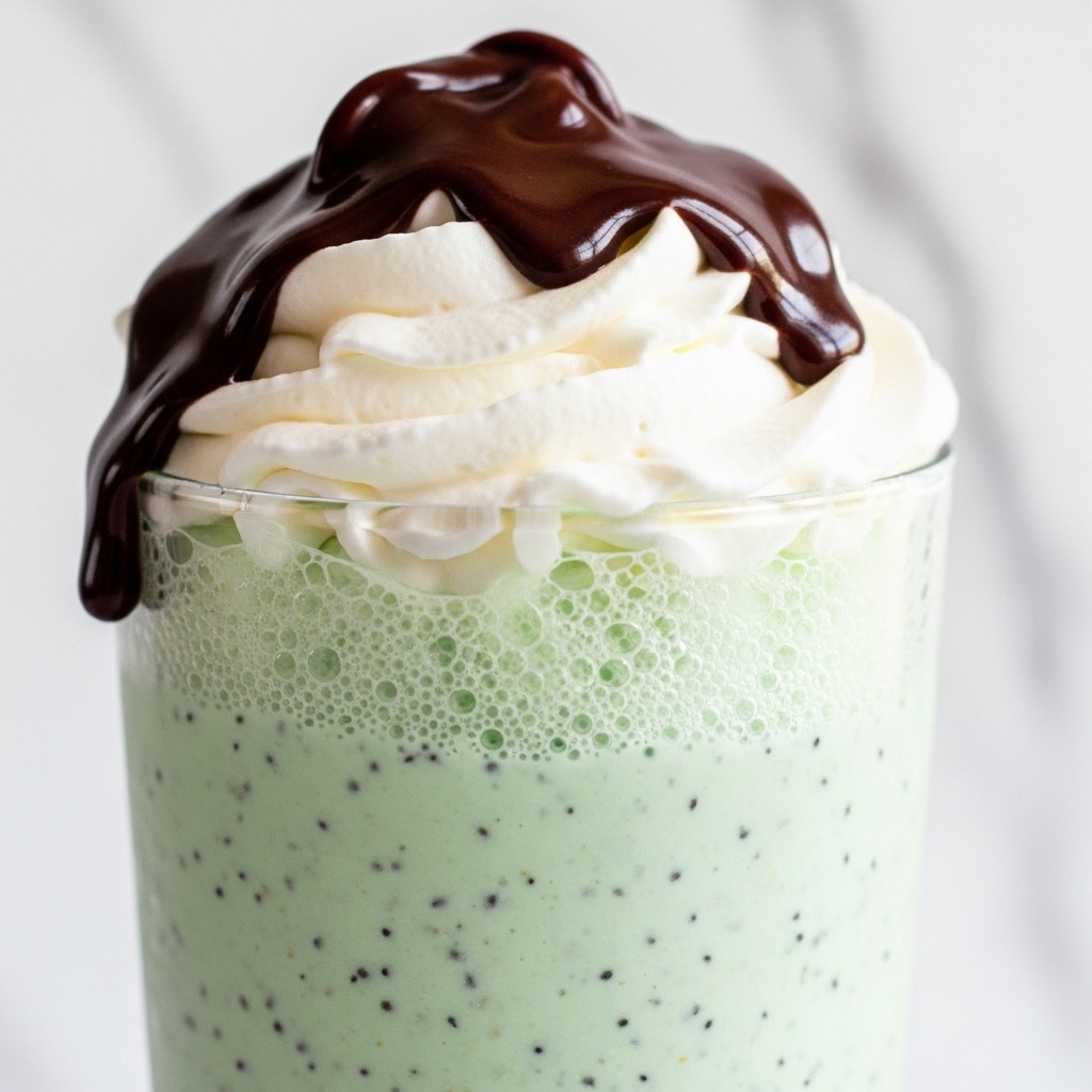 This image shows a close-up of a layered drink in a clear glass. The bottom layer is a light mint green mixture with small dark specks, giving a slightly textured look. Above it is a thin foamy layer with tiny bubbles. The next layer is thick white whipped cream, with a soft and fluffy texture. On top, there is a smooth, thick chocolate sauce dripping down slightly over the whipped cream, dark brown and glossy. The background is a white marbled texture. photo taken with an iphone --ar 4:5 --v 7