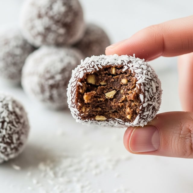 A close-up image of a small round chocolate ball covered in white coconut flakes held between a woman's thumb and forefinger, with a bite taken from it showing a dense, textured dark brown inside with bits of nuts or oats. In the softly blurred background, similar chocolate balls are stacked on a white marbled surface. The focus is on the chocolate ball in the foreground with coconut flakes scattered around it. Photo taken with an iphone --ar 4:5 --v 7