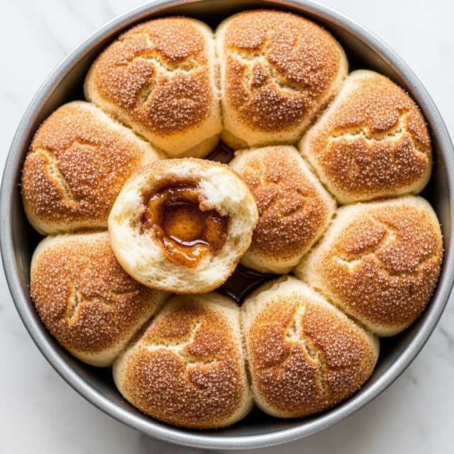 The image shows a round metal pan filled with seven soft, golden-brown biscuits covered in a thick layer of cinnamon sugar, their tops slightly cracked and textured with the sugar coating. One biscuit is broken open, revealing a shiny, sticky cinnamon syrup filling inside with a soft, fluffy interior around it. The biscuits are closely packed together, resting on the white marbled surface beneath the pan. The cinnamon sugar adds a textured, gritty look on top of the biscuits, while the syrup inside looks glossy and sticky. photo taken with an iphone --ar 4:5 --v 7