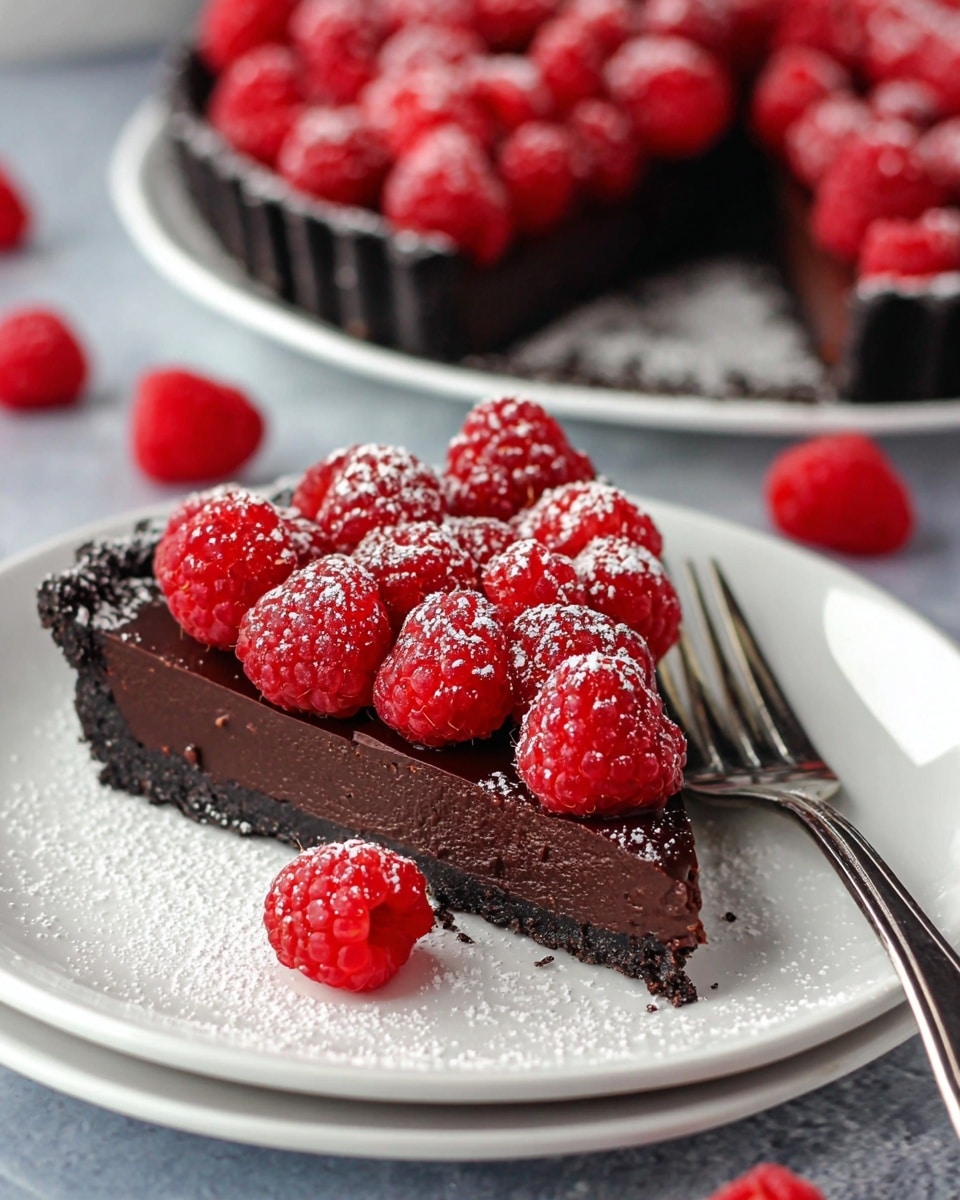 A slice of rich dark chocolate tart with a black crumbly crust, topped with a pile of bright red fresh raspberries that are dusted lightly with powdered sugar. The tart slice lies on a white plate with a shiny finish, alongside a silver fork resting on the right side of the plate. More raspberries and another full tart are seen blurred in the background on a white marbled texture. Powdered sugar is scattered lightly around the plate and raspberries. Photo taken with an iphone --ar 4:5 --v 7