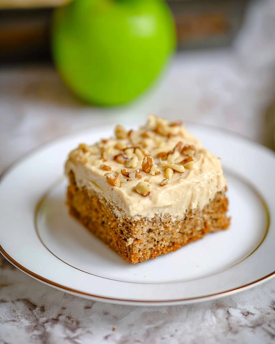 A single square piece of brown cake with a soft texture sits centered on a white plate with two thin gold rings near the rim. The cake has a thick layer of creamy light beige frosting on top, sprinkled with small pieces of chopped walnuts. In the background, there is a blurred bright green apple on a white marbled textured surface. Photo taken with an iphone --ar 4:5 --v 7