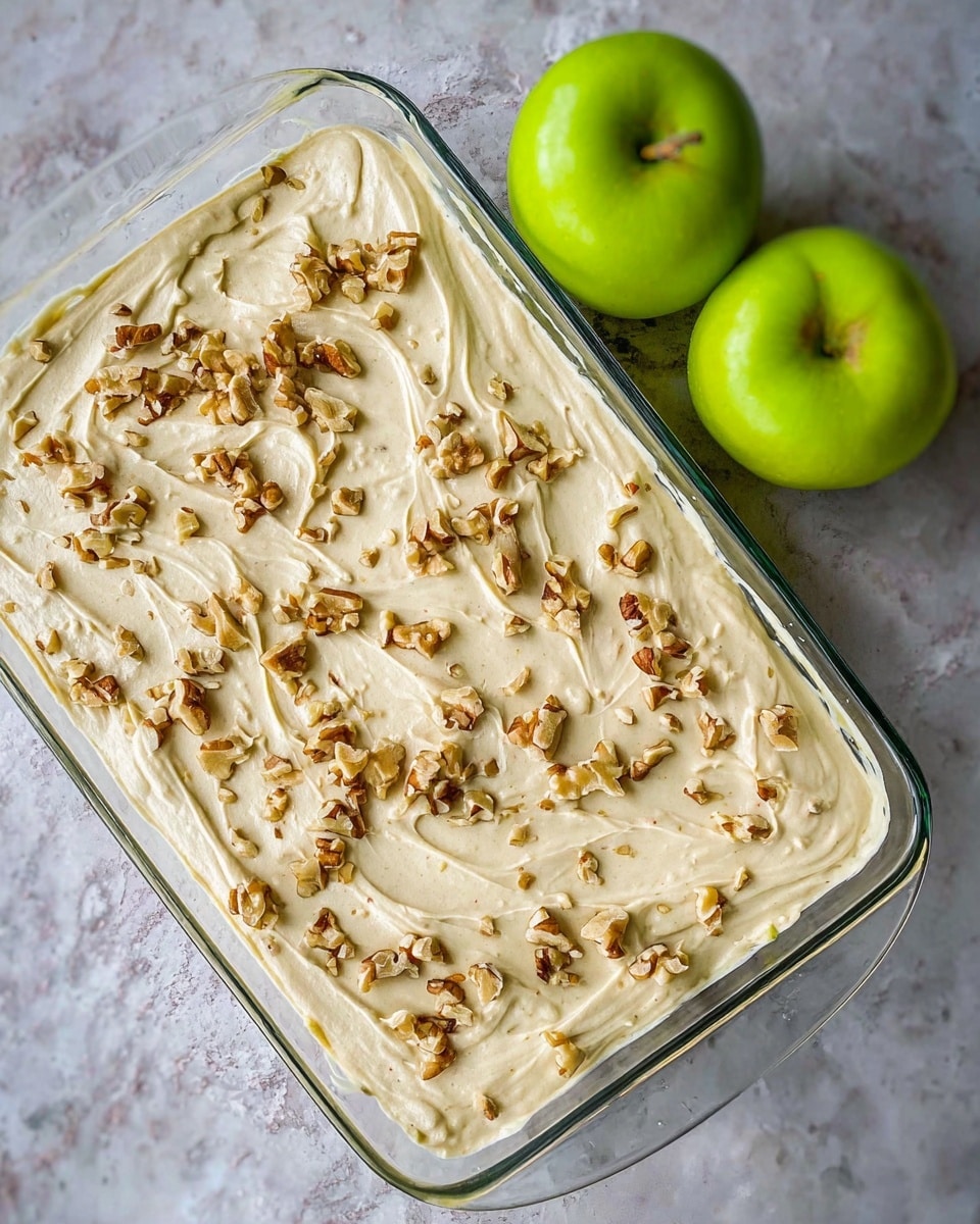 A clear glass rectangular dish holds a creamy beige batter that fills the dish and is spread smoothly with soft waves and swirls on the surface. Small chopped walnut pieces are scattered evenly on top, adding a crunchy texture and a mix of light and dark brown colors. The dish is placed on a white marbled textured surface. Two bright green apples, shiny and smooth, are positioned close to the top right corner of the dish. photo taken with an iphone --ar 4:5 --v 7