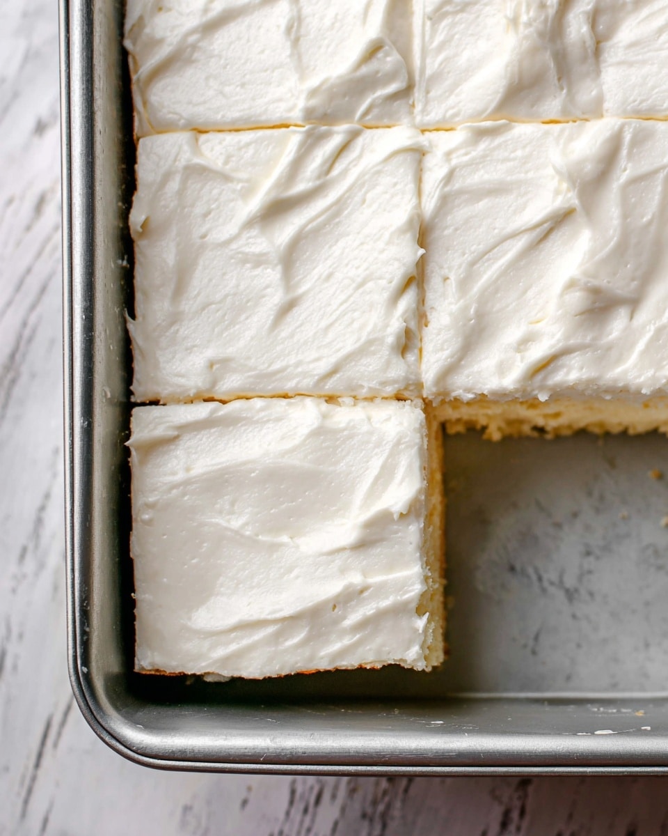 The image shows a close-up view of a rectangular dessert cut into square pieces inside a silver baking pan. The dessert has two visible layers: the bottom layer is light golden brown, firm and cake-like, while the top layer is thick and creamy with a smooth, slightly textured white frosting spread evenly over the surface. The frosting covers the entire top of the dessert, creating a soft contrast with the golden base beneath it. The pan is set on a white marbled surface, and two square pieces are neatly cut but left inside the pan, showing clean edges between them. photo taken with an iphone --ar 4:5 --v 7