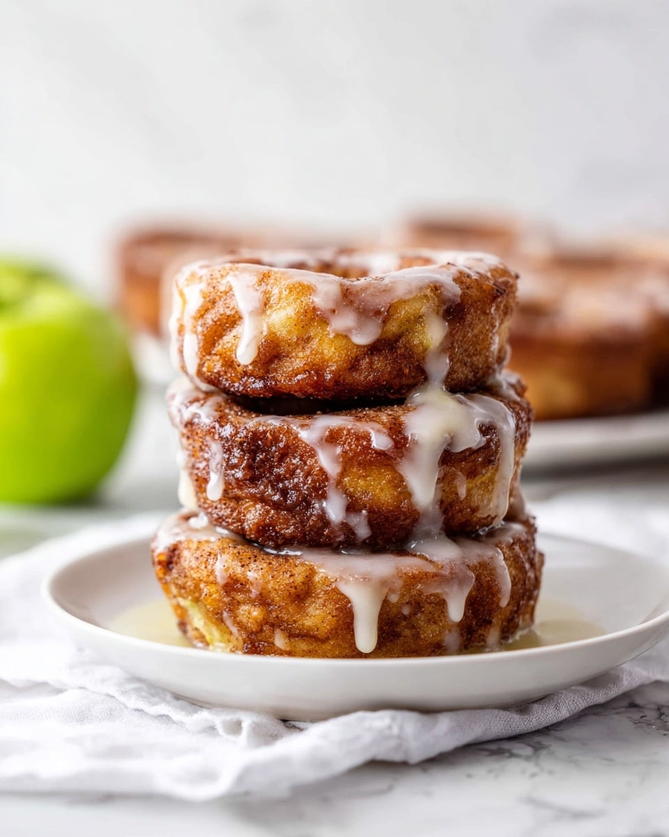The image shows a stack of three thick, round cinnamon rolls on a white plate, placed on a white cloth with a white marbled background. Each cinnamon roll has a golden-brown, crispy outer layer with a soft, textured inside, and they are all generously drizzled with a smooth white glaze that drips down the sides. The top roll is slightly tilted, revealing the gooey layers inside, while the middle roll is darker and more caramelized. In the blurred background, there are more cinnamon rolls and a green apple visible on the left side. Photo taken with an iphone --ar 4:5 --v 7