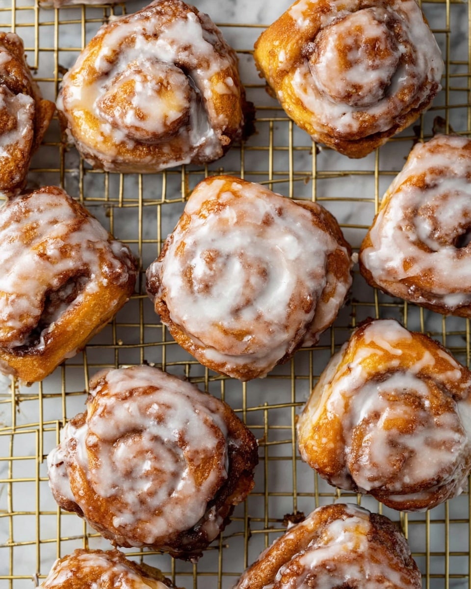 A close-up view of eleven irregularly shaped cinnamon rolls arranged on a gold cooling rack over a white marbled textured surface, each roll showing visible layers of golden-brown dough swirled with darker cinnamon filling and topped with a shiny, uneven glaze of white icing that pools slightly in creases and highlights the texture; the rolls vary slightly in size and shape, with some edges more rounded and others more jagged, emphasizing a fresh, homemade look. photo taken with an iphone --ar 4:5 --v 7