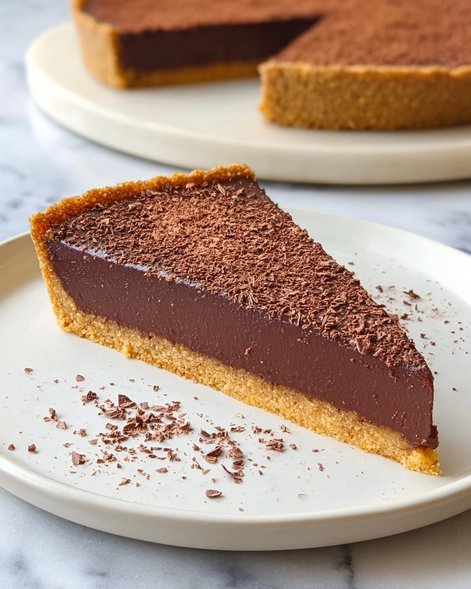 A single slice of chocolate tart sits on a white plate, showing three clear layers: a thin, rough-textured light brown crumb crust at the bottom and edges; a thick, smooth dark brown chocolate filling in the middle with a creamy finish; and a top layer of finely grated chocolate sprinkled evenly across, adding a slightly rough texture. The plate itself has small chocolate flakes scattered around the slice. In the background, the full tart with the same three layers is visible, resting on a white marbled surface. photo taken with an iphone --ar 4:5 --v 7