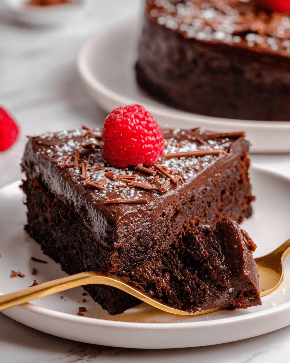 The image shows a close-up of a thick slice of dark chocolate cake on a white plate with a smooth, glossy chocolate frosting layer on top, sprinkled lightly with powdered sugar and thin chocolate shavings. A bright red raspberry sits on the top center of the cake, adding a fresh pop of color. Part of the cake slice is cut off and held by a golden fork, showing the dense, moist texture of the chocolate cake inside. Another similarly decorated slice is visible in the background on a white plate, all set on a white marbled surface. photo taken with an iphone --ar 4:5 --v 7