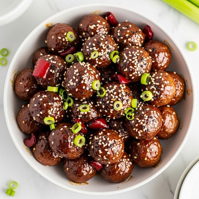 The image shows a white bowl filled with about two layers of shiny, dark brown meatballs covered in a sticky glaze. Mixed among the meatballs are small dark red chunks, likely pieces of red pepper, also glossy from the sauce. The dish is sprinkled with white and black sesame seeds and garnished with thinly sliced green onion rings, which add a fresh bright green color on top. The bowl is set on a white marbled surface, and parts of two other white bowls and a green stalk are visible at the edges. Photo taken with an iphone --ar 4:5 --v 7