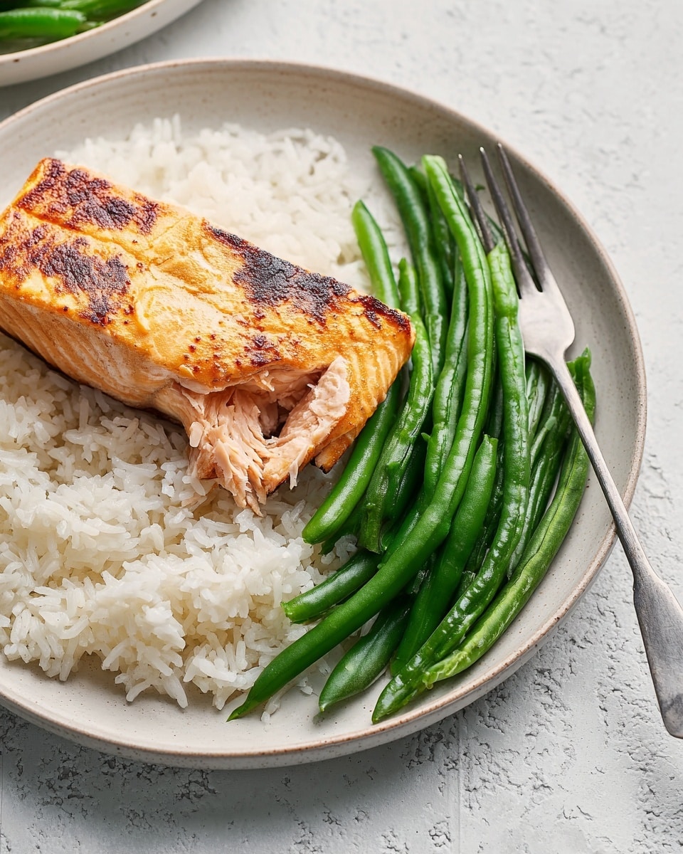 A white round plate holds a serving of three layers: at the bottom, a bed of fluffy white rice with visible individual grains; on top of the rice lies a large piece of cooked salmon with a golden-brown seared surface showing light char marks, with a small broken-off piece revealing the moist pink interior; next to the salmon and rice is a neat pile of bright green cooked green beans with smooth surfaces and some tips visible. A silver fork rests on the right side of the plate, partially on the green beans. The plate sits on a white marbled textured surface. Photo taken with an iphone --ar 4:5 --v 7