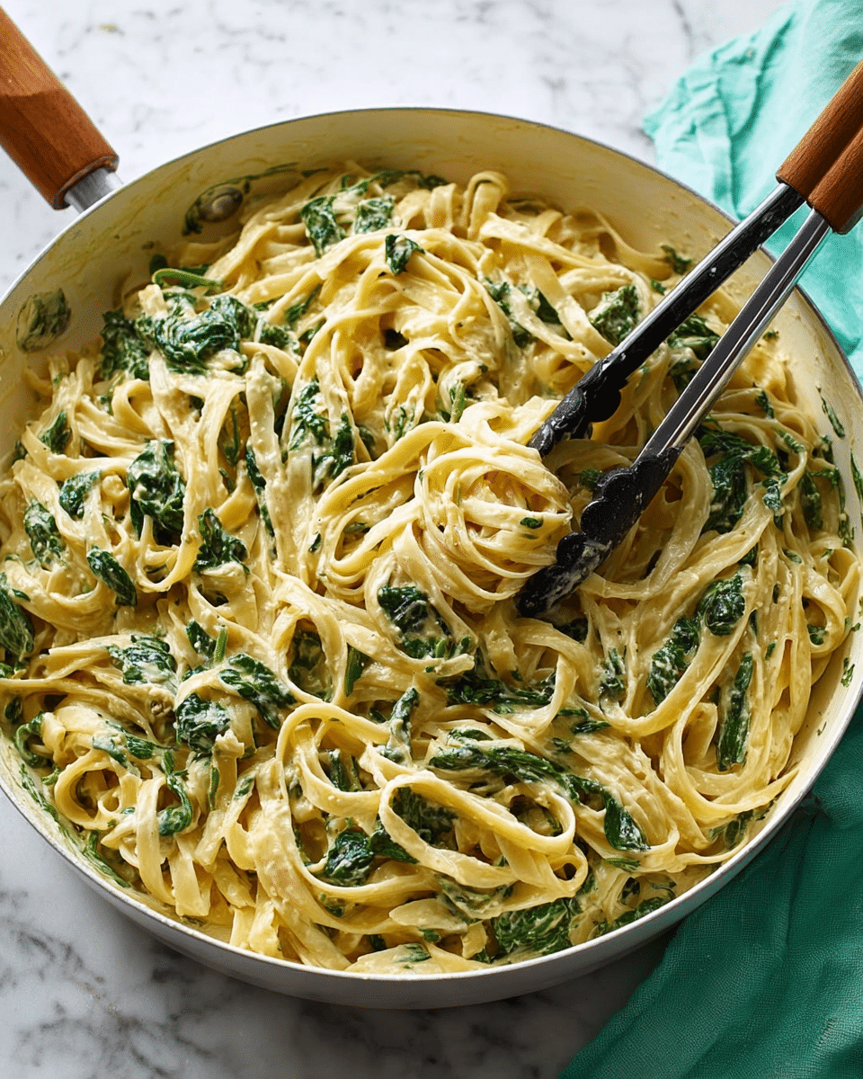 A white pan filled with creamy fettuccine pasta mixed with bright green spinach leaves throughout. The pasta is coated evenly in a smooth, light beige sauce, giving it a shiny and rich texture. Two black metal tongs rest inside the pan, slightly lifting some of the pasta. The pan handle has a wooden brown grip, and the pan sits on a white marbled surface with a mint green cloth partially visible at the edge. photo taken with an iphone --ar 4:5 --v 7