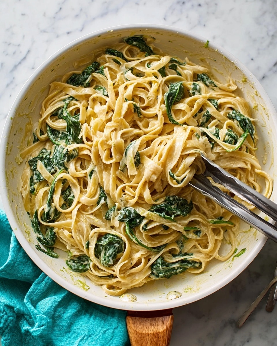 A white frying pan with a wooden handle holds creamy fettuccine pasta mixed with wilted green spinach leaves, coated evenly in a light beige sauce with a smooth texture. Two metal tongs are placed inside the pan, partially lifting some noodles. The pan sits on a white marbled surface with a turquoise cloth partially visible to the right of the handle. photo taken with an iphone --ar 4:5 --v 7