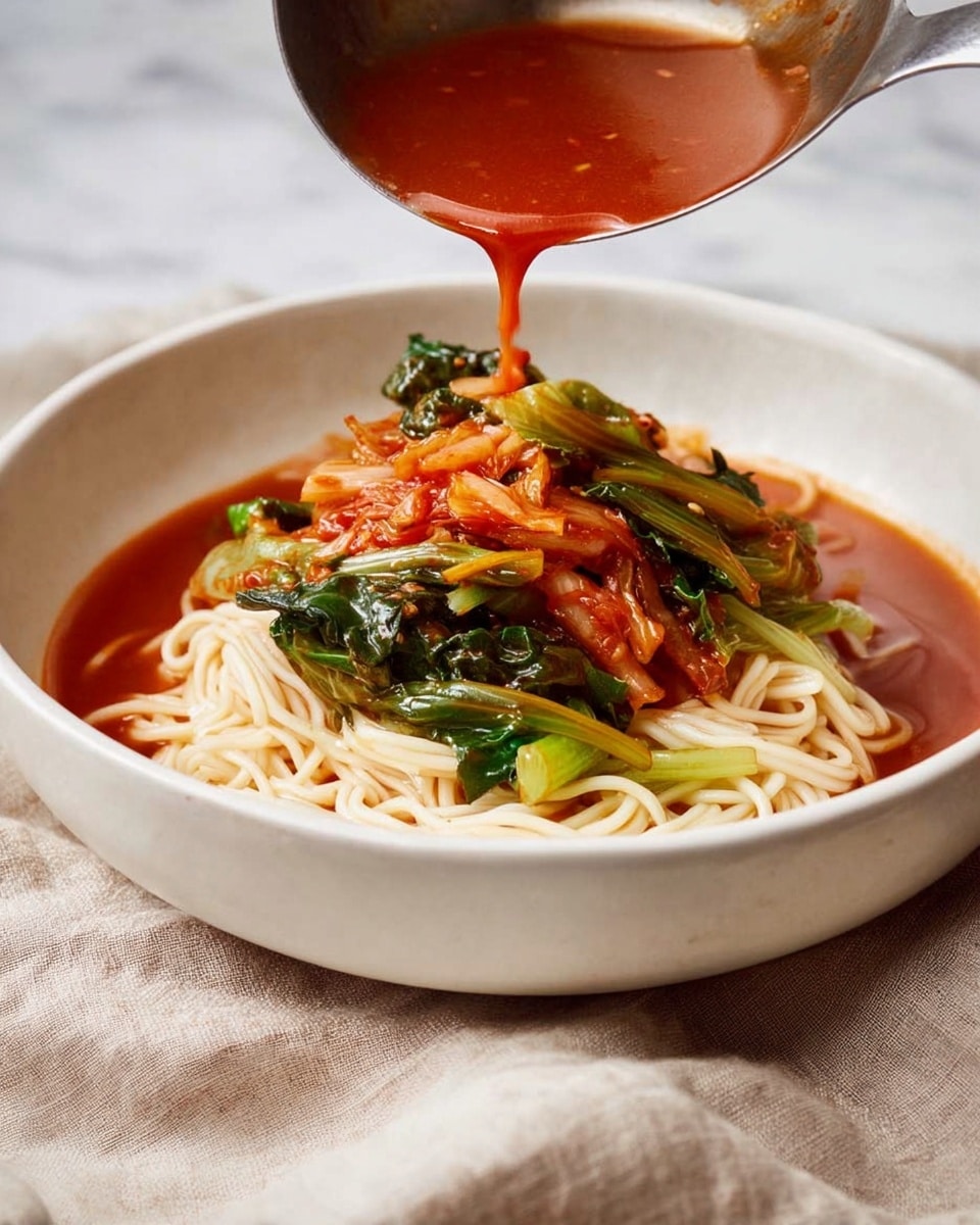 A white bowl filled with three main layers: at the bottom, light cream-colored cooked noodles are neatly arranged, topped by bright green leafy vegetables mixed with reddish pickled cabbage, all covered by a thick bright red broth being poured from a metal ladle above, showing the soup’s glossy and rich texture; the overall scene sits on a white marbled surface with a soft beige cloth beneath the bowl. photo taken with an iphone --ar 4:5 --v 7