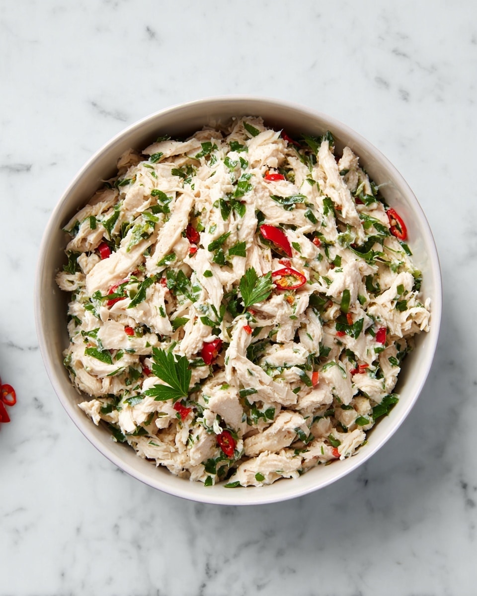 The image shows a bowl of chicken salad in a white bowl, placed on a white marbled surface. The salad consists of shredded white chicken meat as the main layer, mixed with chopped green herbs scattered evenly throughout. There are small pieces of red chili peppers adding color contrast among the chicken and herbs. The texture of the chicken is flaky and moist, while the herbs appear fresh and slightly glossy. The bowl is filled nearly to the top, showing a rich combination of colors and textures. The photo taken with an iphone --ar 4:5 --v 7